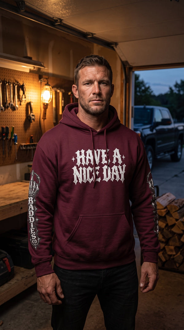 Rugged male model in a garage wearing a maroon Have a Nice Day Bad Weather Baddies graphic hoodie.