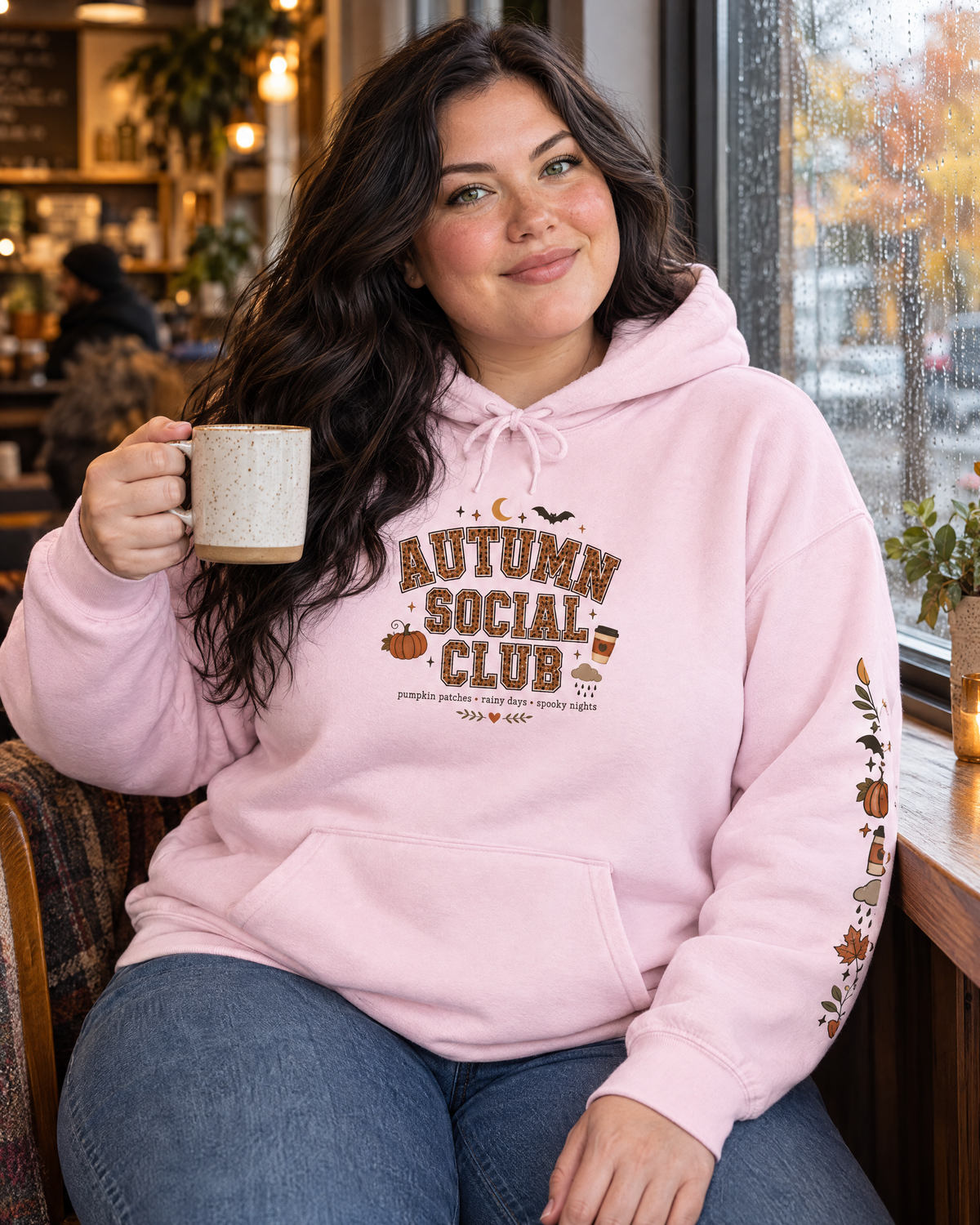 Autumn Social Club hoodie in light pink on a plus-size model in a cozy rainy coffee shop