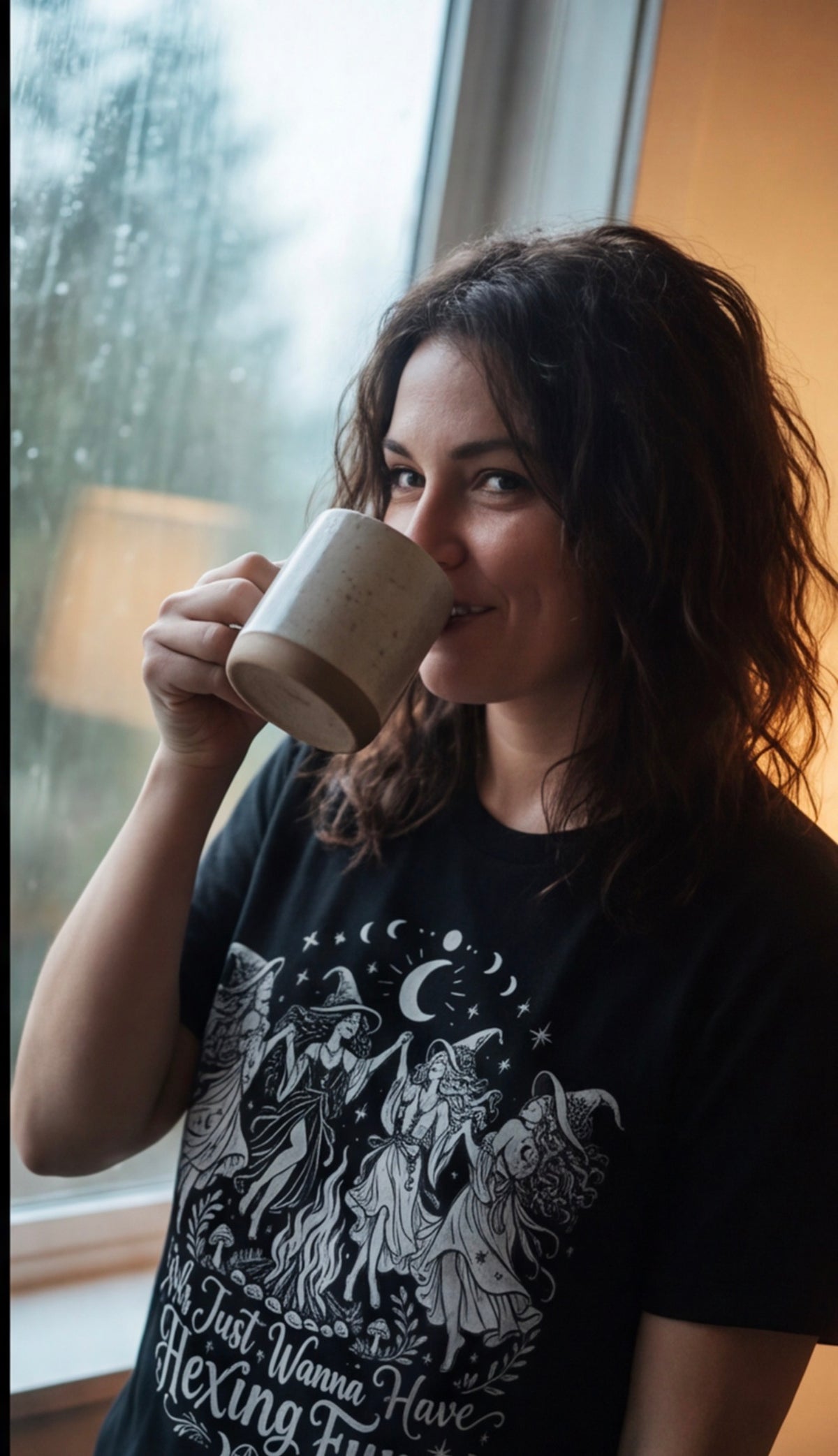 Woman drinking coffee by a rain-streaked window in the Girls Just Wanna Have Hexing Fun witch tee.