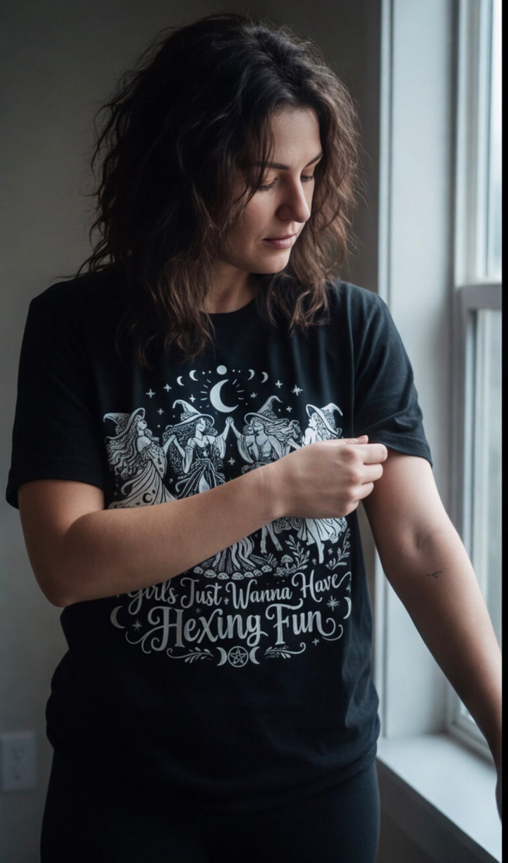 Woman wearing the Girls Just Wanna Have Hexing Fun witch tee beside a rainy window.