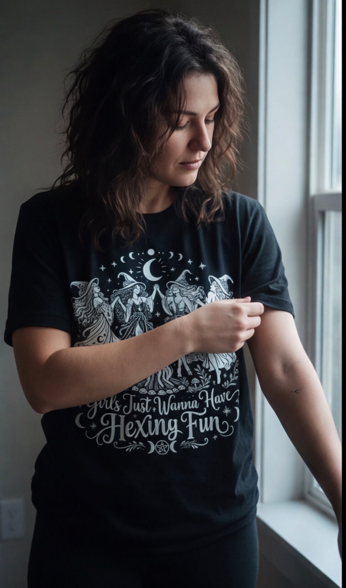 Woman wearing the Girls Just Wanna Have Hexing Fun witch tee beside a rainy window.